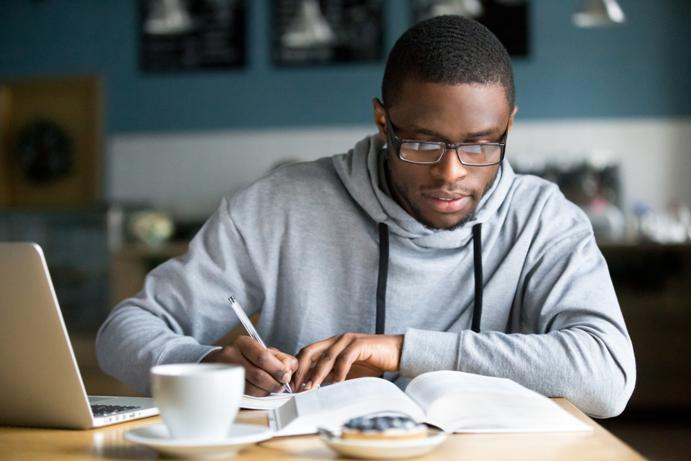 Young male student with glasses sitting at a café table with his laptop and writing in a notebook