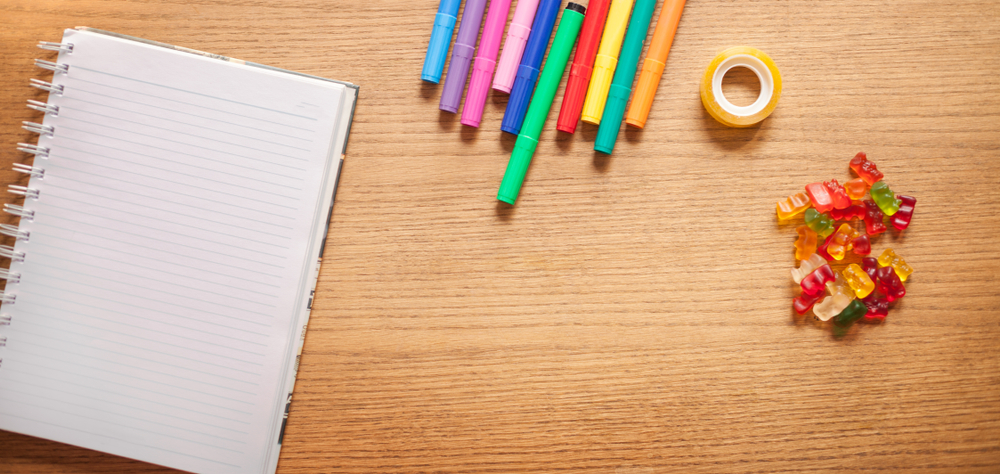 A desk with colorful gummy bears, tape, multicolored pens, and a spiral notebook