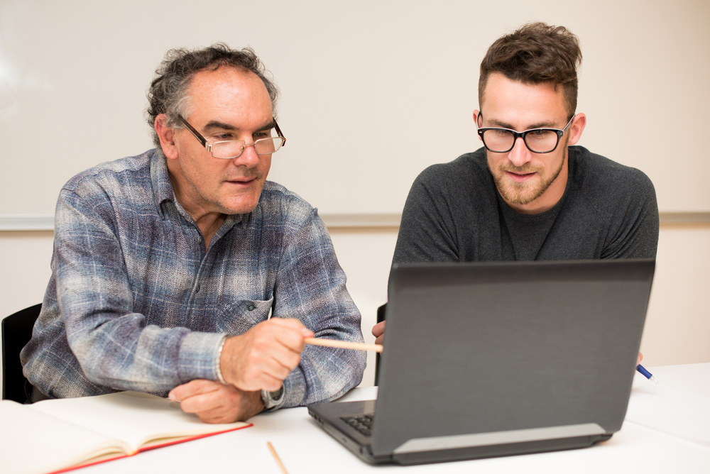 A young male student with glasses sitting at his laptop with his mentor who is pointing to something on the computer screen